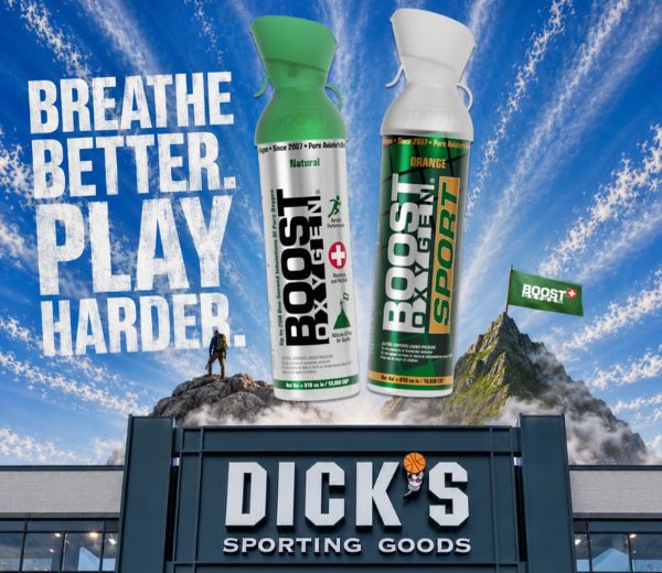 Two Boost Oxygen cans, one Natural and one Sport, are shown floating above a building sign reading 'DICK'S SPORTING GOODS' against a dramatic blue sky with clouds and a mountain. Large text on the left reads 'BREATHE BETTER. PLAY HARDER'