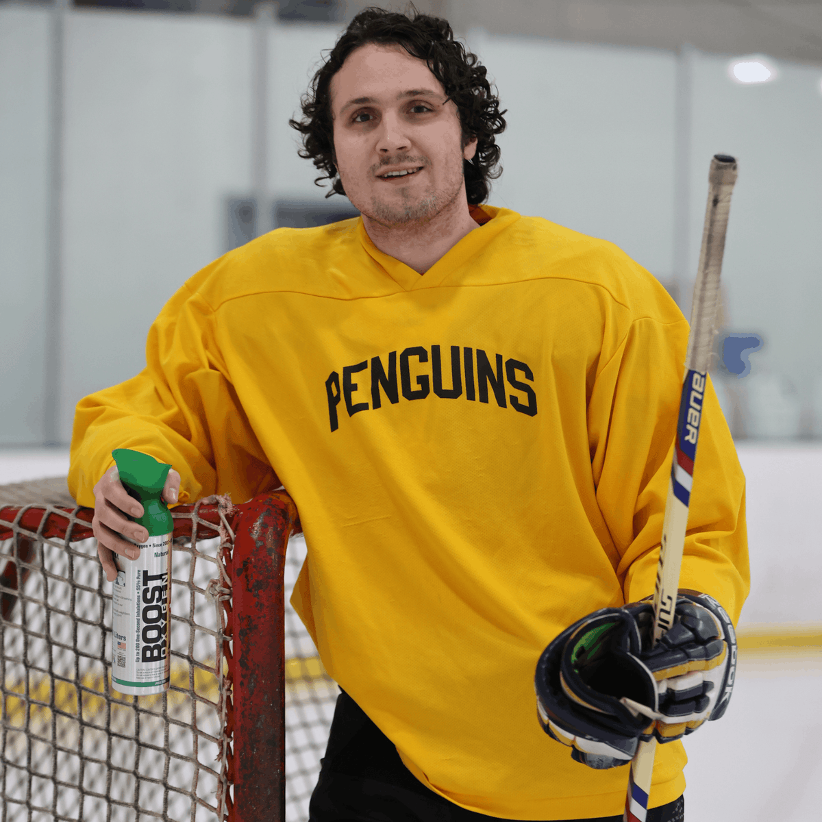 Person wearing a yellow 'PENGUINS' jersey holding a hockey stick and a can of Boost Oxygen on an ice rink.