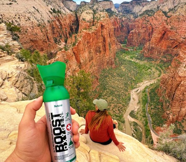 A hand holds a can of Boost Oxygen Natural in front of a scenic red rock canyon with a woman sitting on a cliff edge overlooking a winding river valley.