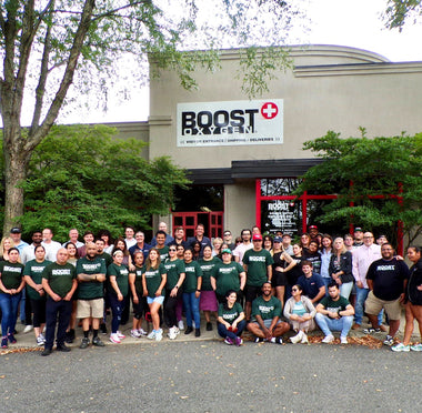 Boost Oxygen staff posing together in front of their building with a prominent 'BOOST SYSTEM' sign. Many individuals are wearing matching dark green t-shirts with white text, while others wear casual clothing.