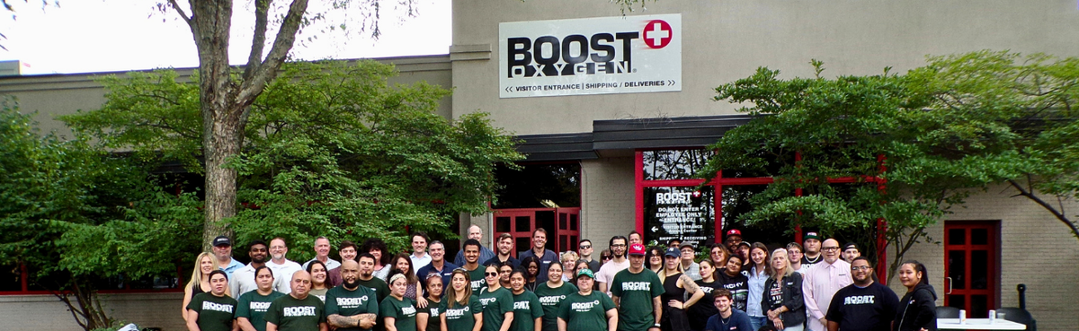 Boost Oxygen employees posing in front of their building with a prominent Boost Oxygen sign in the background. Many individuals are wearing dark green t-shirts with the Boost Oxygen logo.